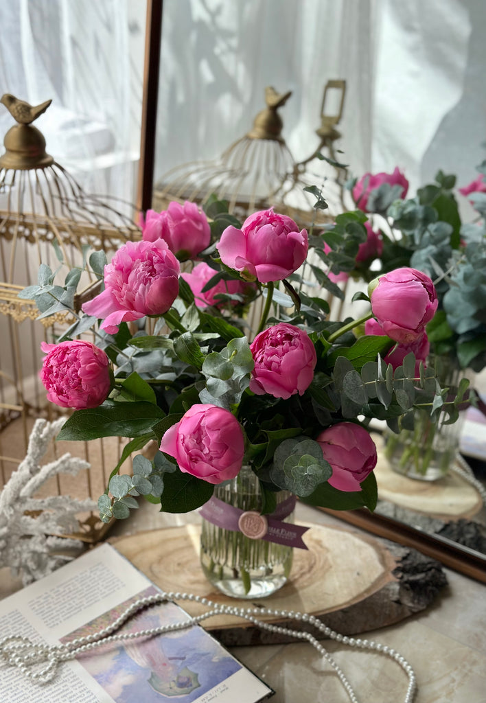 Pink peony flower arrangement in a glass vase on a slab of wood, surrounded by decor