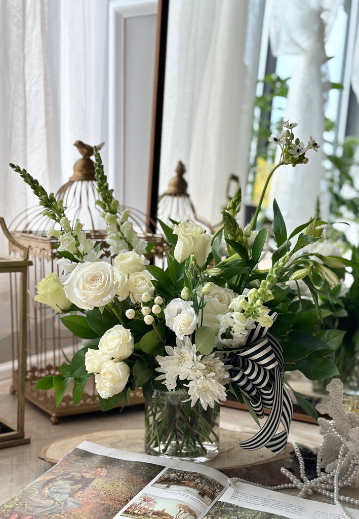 Photo of the Peace Sympathy Arrangement with white flowers in a glass vase surrounded by decor