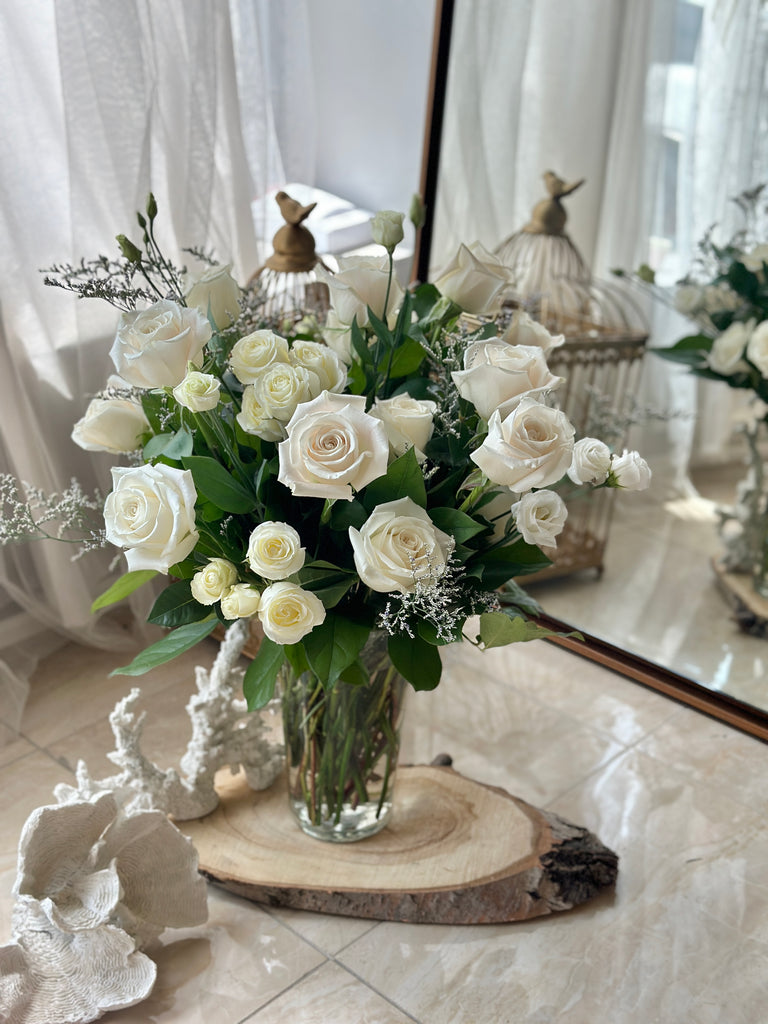 Photo of white roses in a glass vase on a wood slab surrounded by decor